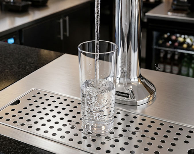 Close-up of a polished chrome beverage tap dispensing water into a clear tumbler positioned on a perforated stainless steel drip tray. Bubbles are visible in the glass as it fills.