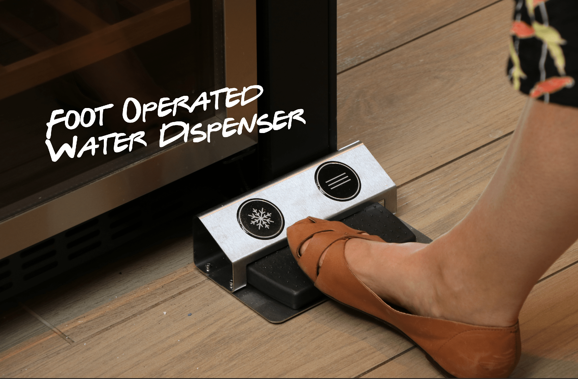 Foot in a brown shoe pushing on a black foot pedal on a silver bracket with a snowflake badge on a laminate wooden floor with the text 'foot operated water dispenser' on the left hand side