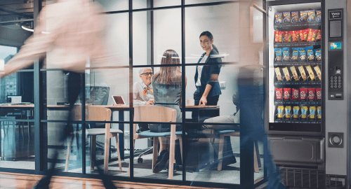 three people in a meeting room while people are walking around by a vending machine